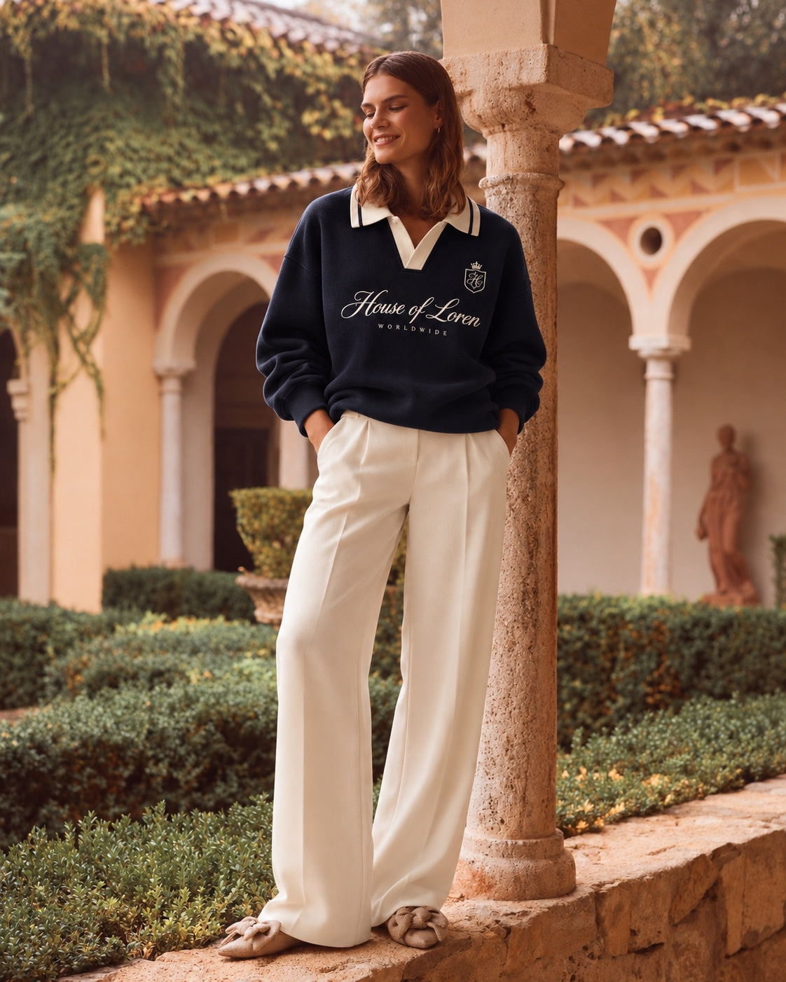 House of Loren – Model im dunkelblauen Pullover mit cremefarbener Hose vor mediterraner Architektur